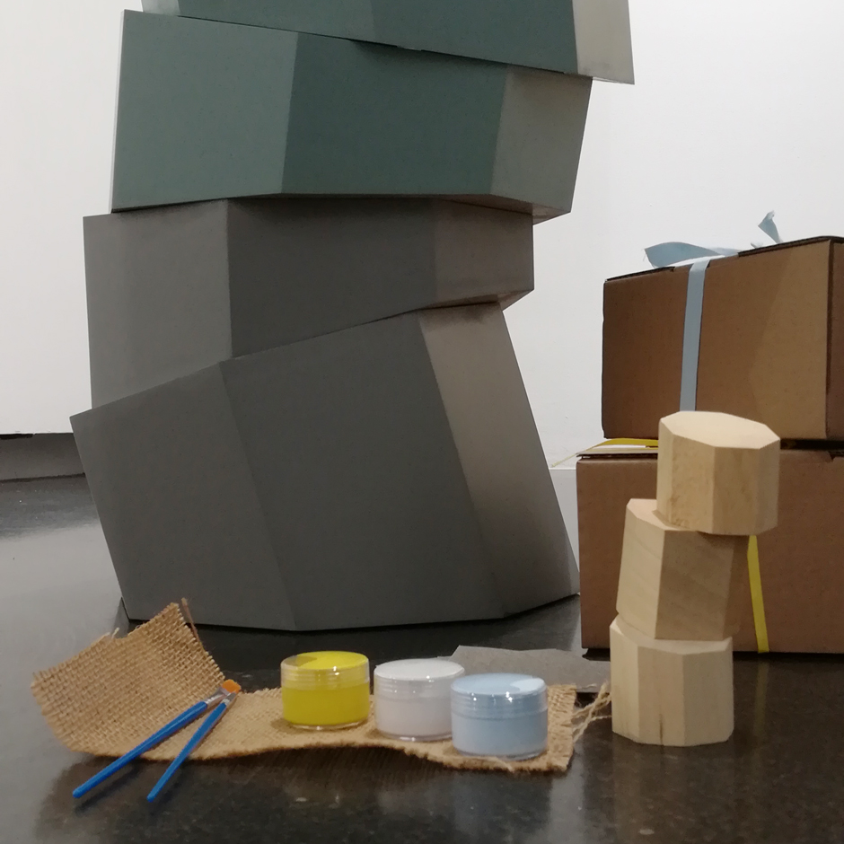 Vista detalle de la obra 'Columna Platónica'. Frente a ella, en el suelo, dos cajas de cartón, apiladas y decoradas, y el contenido del paquete 'Arte' en Familia.